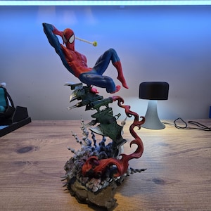 May include: A red and blue Spider-Man action figure is posed in a dynamic action pose. The figure is perched atop a green and gray spaceship-like base with red flames erupting from the base.