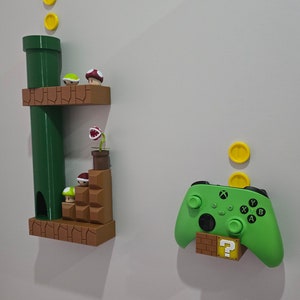 Może przedstawiać: Zielony kontroler do gier wideo z blokiem z pytajnikiem i brązową cegłą pod spodem. Kontroler jest zamontowany na białej ścianie. Po lewej stronie obrazu znajdują się trzy żółte monety nad zieloną rurą i brązową platformą z czerwonym i zielonym grzybem.