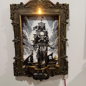 Könnte beinhalten: Gerahmte Piratenschiff-Szene mit Totenkopf-Flagge. Der Rahmen ist verziert mit goldfarbenem Finish und kleinem Licht. Das Schiff ist schwarz mit weißen Segeln, vor einem stürmischen, grauen Himmel. Der Schriftzug "Black Pearl" befindet sich unten.