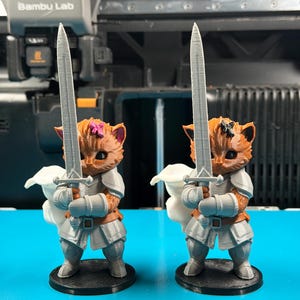 Op de afbeelding: Twee 3D-geprinte oranje kattenbeeldjes in zilveren harnas met lange zwaarden. De ene kat heeft een roze strik op het hoofd, de andere een zwarte. Beide figuren staan op ronde zwarte voetstukken voor een Bambu Lab 3D-printer.