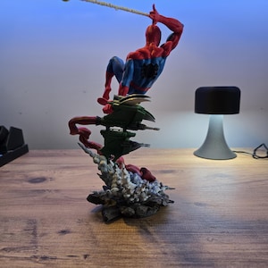 May include: A red and blue Spider-Man action figure is posed in mid-air, swinging from a web. The figure is perched on a green and gray base with a red and gray explosion effect.