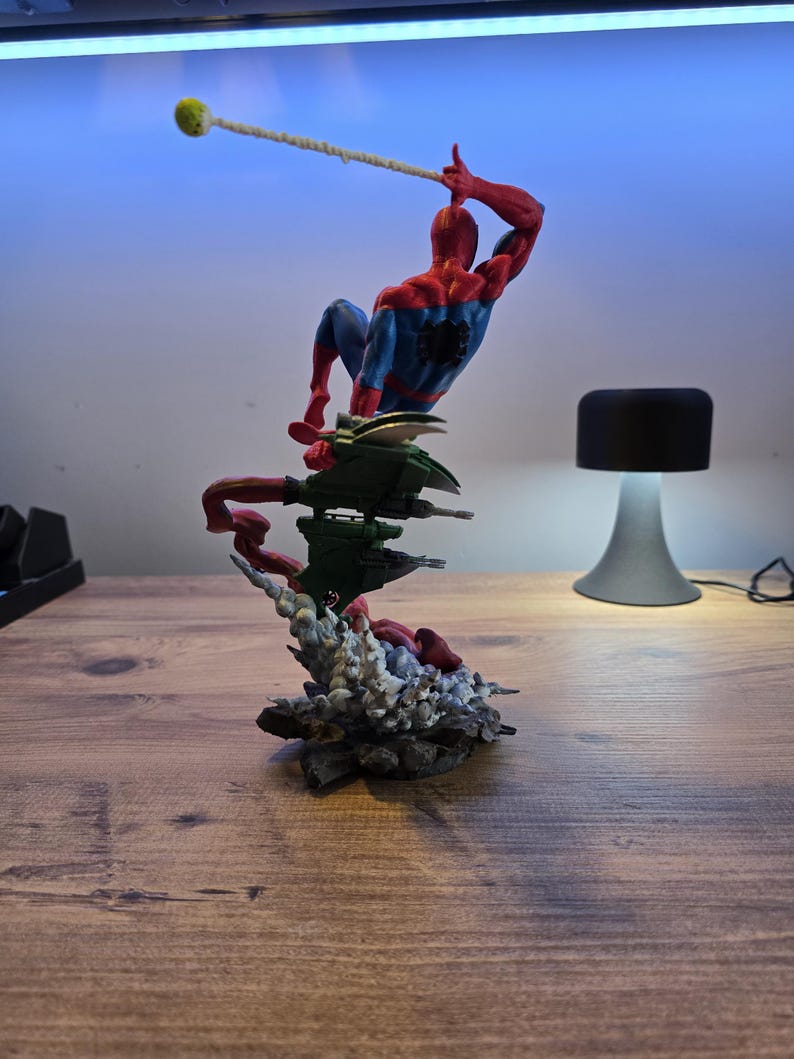 May include: A red and blue Spider-Man action figure is posed in mid-air, swinging from a web. The figure is perched on a green and gray base with a red and gray explosion effect.