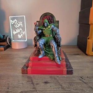 May include: A detailed figurine of a character in a green hooded cloak and silver armor, seated on an ornate throne with a red carpeted base. The throne is brown and gold. The figurine is a collectible item.