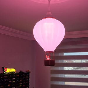 Könnte beinhalten: Eine rosa Deckenleuchte in Form eines Heißluftballons. Die Leuchte hat einen weißen oberen Teil und einen Korb mit einer Hasenfigur. Das Licht hängt von der Decke in einem Raum mit einer Sammlung von Spielzeugautos.