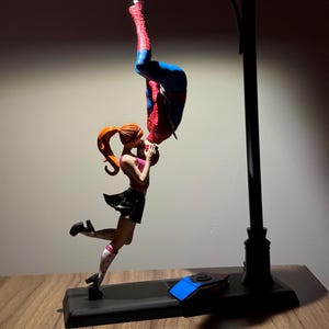 Peut inclure: Une lampe repr&eacute;sentant une figurine de Spiderman suspendue &agrave; l'envers, embrassant une figurine f&eacute;minine aux cheveux roux. La lampe a une base noire et un design de lampadaire. Les figurines sont aux couleurs vives, la lampe diffusant une lueur chaleureuse.