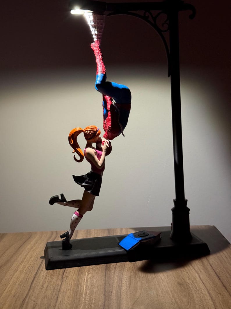 Peut inclure: Une lampe repr&eacute;sentant une figurine de Spiderman suspendue &agrave; l'envers, embrassant une figurine f&eacute;minine aux cheveux roux. La lampe a une base noire et un design de lampadaire. Les figurines sont aux couleurs vives, la lampe diffusant une lueur chaleureuse.