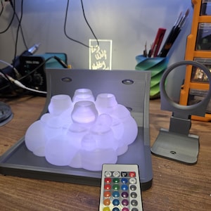 May include: A glowing, cloud-shaped lamp with a remote control. The lamp is composed of multiple translucent, rounded forms, emitting a soft, diffused light. The remote control has various color options. The lamp sits on a gray base.