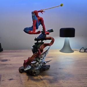 May include: A red and blue Spider-Man action figure is posed in a dynamic action pose, swinging from a web line. The figure is perched on a detailed base with a red and black smoke effect.