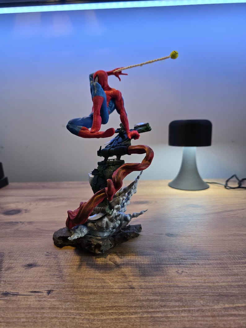 May include: A red and blue Spider-Man action figure is posed in a dynamic action pose, swinging from a web line. The figure is perched on a detailed base with a red and black smoke effect.