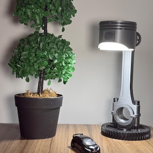 Könnte beinhalten: Ein schwarzes Spielzeugauto steht auf einem Holztisch neben einer Lampe aus einem Auto-Kolben. Im Hintergrund steht eine Topfpflanze.