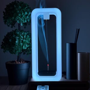 May include: A white LED incense burner with a rectangular frame and a remote control. The incense stick is burning and smoke is rising inside the frame. The burner is on a wooden base and there are two small potted trees in the background.