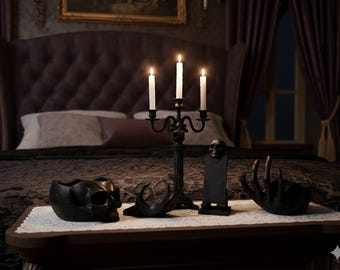 May include: A gothic bedroom scene with a black candelabra holding three lit candles, skull-shaped ornaments, and a decorative hand. The room features a large, buttoned headboard and dark curtains.