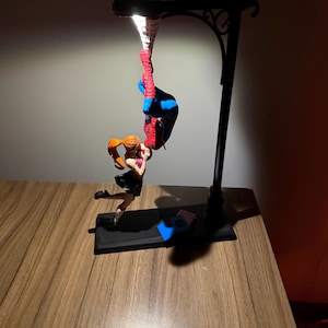 Peut inclure: Un lampadaire noir avec une figurine de Spiderman suspendue &agrave; l'envers. Une figurine de femme aux cheveux roux tend la main vers Spiderman. Le lampadaire est sur une surface en bois.