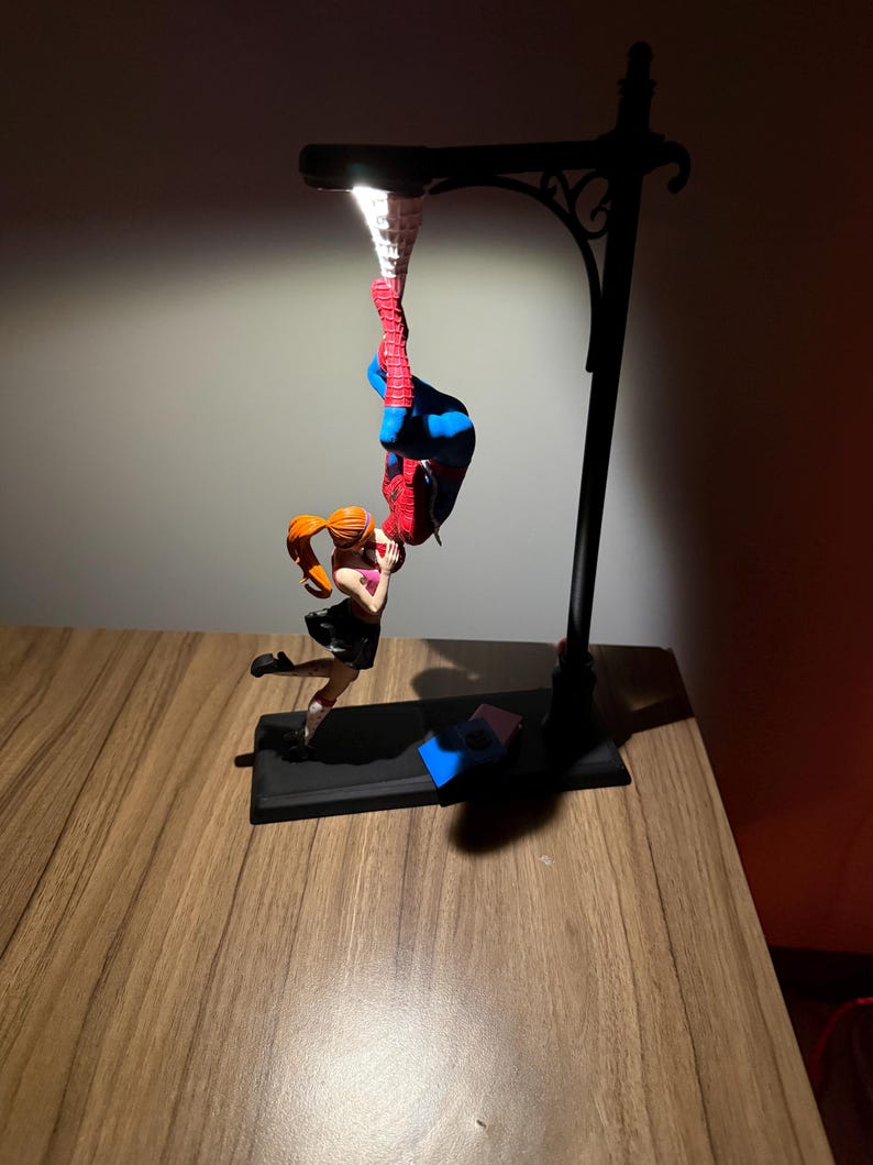 Peut inclure: Un lampadaire noir avec une figurine de Spiderman suspendue &agrave; l'envers. Une figurine de femme aux cheveux roux tend la main vers Spiderman. Le lampadaire est sur une surface en bois.