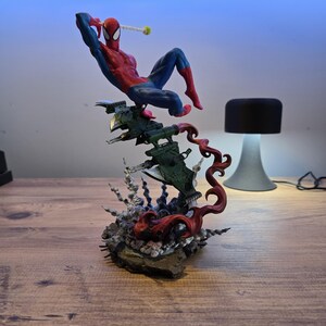 May include: A red and blue Spider-Man action figure is posed in a dynamic action pose, perched atop a green and black spaceship-like base. The base is surrounded by red, black, and white tendrils, creating a dramatic and dynamic scene.