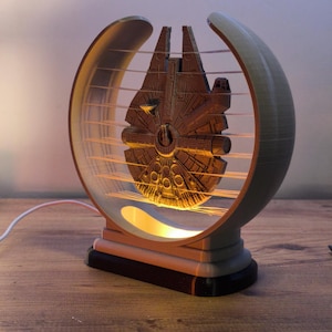 Könnte beinhalten: Eine 3D-gedruckte Lampe mit einem detaillierten Modell des Raumschiffs Millennium Falcon. Die Lampe ist in einem kreisförmigen Rahmen mit horizontalen Linien eingefasst und strahlt ein warmes, goldenes Licht aus.