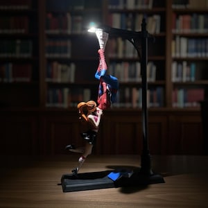 Peut inclure: Lampe d&eacute;corative repr&eacute;sentant Spiderman suspendu la t&ecirc;te en bas, avec une figurine de femme tendant les bras vers lui. La lampe a une base noire et un design de lampadaire, avec une lumi&egrave;re chaude &eacute;clairant les figurines. En arri&egrave;re-plan, une biblioth&egrave;que.