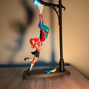 Peut inclure: Lampe d&eacute;corative repr&eacute;sentant un super-h&eacute;ros suspendu &agrave; l'envers &agrave; un lampadaire. Une figurine f&eacute;minine en uniforme scolaire tend la main vers le super-h&eacute;ros. La lampe a une base noire et une lumi&egrave;re bleue.