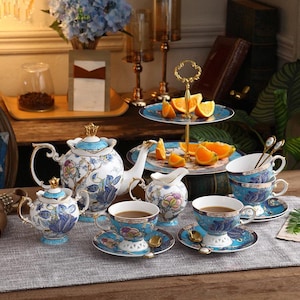 Könnte beinhalten: Ein komplettes Porzellan-Teeservice mit blau-weißem Blumendesign. Das Set beinhaltet eine Teekanne, einen Sahnekännchen, eine Zuckerdose, Teetassen, Untertassen und Löffel. Ein zweistöckiger Servierständer hält Orangenscheiben. Das Teeservice steht auf einer grauen Tischdecke.