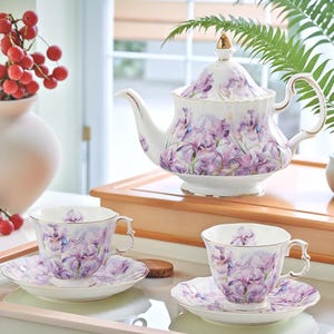 May include: A white teapot and two teacups with matching saucers. The teapot and cups are decorated with a floral pattern of purple irises. The teapot has a gold lid knob and a gold rim. The cups and saucers have gold rims.