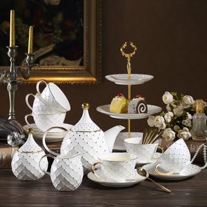 May include: A sophisticated white porcelain tea set with gold detailing and a diamond pattern. The set includes a teapot, creamer, sugar bowl, cups, saucers, and a tiered serving tray. Gold dots and handles adorn the set.