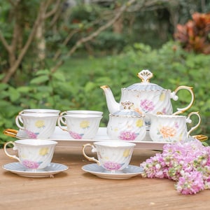 Pode incluir: Um conjunto de chá de porcelana branca com desenhos florais, incluindo uma chaleira, açucareiro, cremeira e seis xícaras com pires. O conjunto tem detalhes dourados e está disposto em uma bandeja branca. Flores rosas e amarelas.