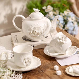 May include: A white ceramic tea set featuring a teapot, two teacups, and saucers. The teapot has a lid with a butterfly-shaped handle and a floral design. The teacups and saucers also have floral details. The set is on a wooden surface.