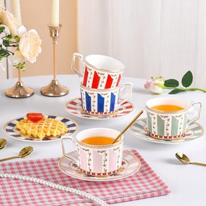 Puede incluir: Juego de tazas y platillos decorativos con diseños florales y a rayas. Las tazas contienen un líquido dorado. Un gofre con fresa está sobre un plato. También hay cucharas y candelabros dorados.
