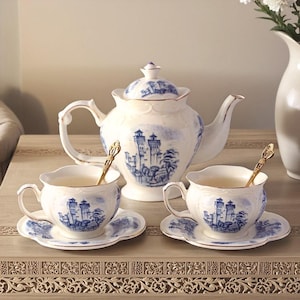 May include: A white porcelain tea set with blue landscape designs. The set includes a teapot, two teacups, saucers, and spoons. The teacups and saucers are on a decorative table. The teapot has a lid and a curved handle.