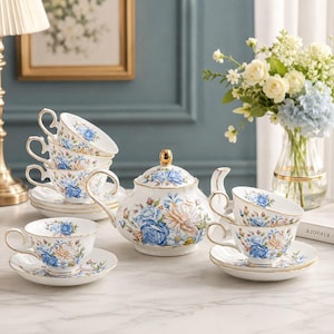 May include: A porcelain tea set featuring a white base with blue and peach rose floral patterns and gold trim. The set includes a teapot with a gold lid knob, multiple teacups with matching saucers, and a stack of saucers, all decorated with roses.