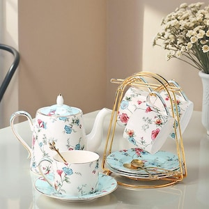 Könnte beinhalten: Weißes Porzellan-Teeservice mit floralem Muster in Rosa, Blau und Grün. Das Set beinhaltet eine Teekanne, Tassen, Untertassen und einen goldfarbenen Metallständer. Die Tassen und Untertassen sind auf einem Tisch ausgestellt.
