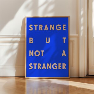 May include: A framed print with a blue background and gold text that reads "STRANGE BUT NOT A STRANGER".