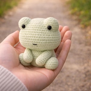 Puede incluir: Una rana de ganchillo verde claro con ojos negros y una boca pequeña. La rana está sentada en una mano. La rana es un artículo hecho a mano.