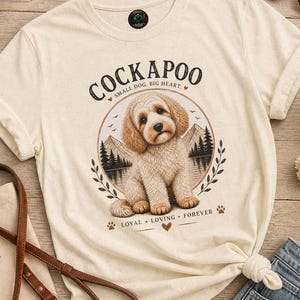 Peut inclure: Un t-shirt couleur crème avec un motif représentant un chien Cockapoo assis devant un paysage de montagnes et de forêt. Le texte indique "COCKAPOO", "SMALL DOG, BIG HEART." et "LOYAL LOVING FOREVER" avec des empreintes de pattes et des cœurs.