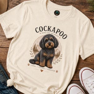 Puede incluir: Una camiseta de manga corta color crema con un gráfico de un perro Cockapoo negro y fuego. El texto dice COCKAPOO, SMALL DOG, BIG HEART y LOYAL, LOVING, FOREVER. El diseño incluye un fondo circular con árboles y montañas.