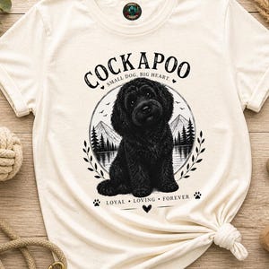May include: A cream-coloured short-sleeve t-shirt featuring a black graphic of a Cockapoo dog sitting in front of a mountain and lake landscape. The text reads COCKAPOO, SMALL DOG, BIG HEART, and LOYAL, LOVING, FOREVER, with small paw print accents.