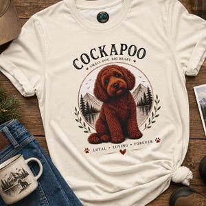 Puede incluir: Una camiseta de color crema con un gráfico de un perro Cockapoo marrón sentado frente a un paisaje de montaña y lago. El texto dice COCKAPOO, SMALL DOG, BIG HEART y LOYAL, LOVING, FOREVER, acompañado de pequeñas huellas y corazones.