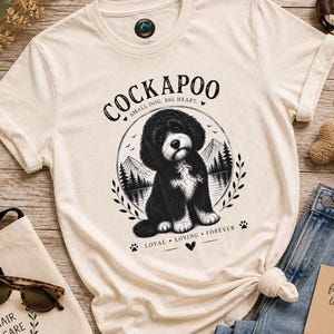 Puede incluir: Una camiseta de manga corta color crema con un gráfico en blanco y negro de un perro Cockapoo. El texto dice COCKAPOO, SMALL DOG, BIG HEART y LOYAL, LOVING, FOREVER. El diseño incluye un marco circular con montañas y árboles.