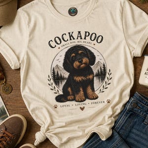 Puede incluir: Una camiseta de color crema con un gráfico de un cachorro Cockapoo negro y fuego sentado frente a un paisaje de montaña y lago. El texto dice "COCKAPOO", "SMALL DOG. BIG HEART." y "LOYAL LOVING FOREVER" con detalles de huellas.