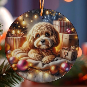 Pode incluir: Enfeite de Natal redondo com um cão castanho claro rodeado de presentes embrulhados e bolas. Luzes de Natal e uma árvore de Natal ao fundo criam um ambiente festivo. O enfeite tem um cordão laranja para pendurar.