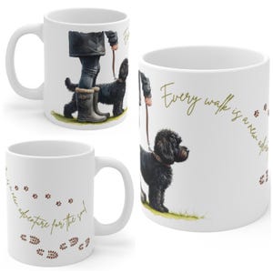 Puede incluir: Taza de cerámica blanca con una ilustración en acuarela de una persona paseando a un perro negro con correa. La taza tiene el texto "Every walk is a new adventure for the soul" en letras doradas, con huellas de patas y de pies.