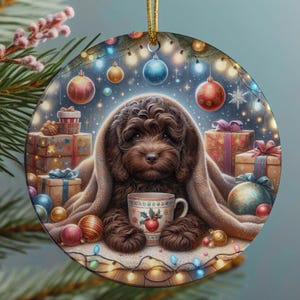 Peut inclure: Une décoration de Noël ronde représentant un chiot brun aux poils bouclés enveloppé dans une couverture beige, tenant une tasse blanche ornée de baies rouges et de feuilles vertes. Le fond montre des cadeaux, des boules et des guirlandes.