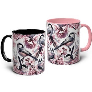Op de afbeelding: Twee keramische mokken met vogel- en bloemmotieven. Eén mok heeft een zwart interieur en handvat, de andere heeft een roze interieur en handvat. De mokken tonen illustraties van vogels die op takken zitten met roze bloesems tegen een witte achtergrond.