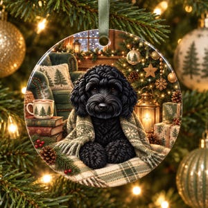 May include: A round ceramic Christmas ornament featuring a black curly-haired dog wrapped in a green and white plaid blanket. The scene includes a mug with pine tree patterns, stacked books, a lit lantern, and wrapped gifts in a cosy room.