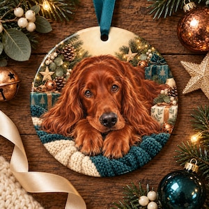 May include: A round ceramic Christmas ornament featuring a detailed illustration of a long-haired Irish Setter dog resting on a textured cream and teal knit blanket, surrounded by festive pine branches, wrapped gifts, and gold star decorations.