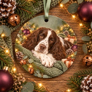 May include: A round ceramic Christmas ornament featuring a brown and white English Springer Spaniel resting on a green knitted blanket, surrounded by pine branches, small pinecones, and wrapped gift boxes with a green ribbon hanger.