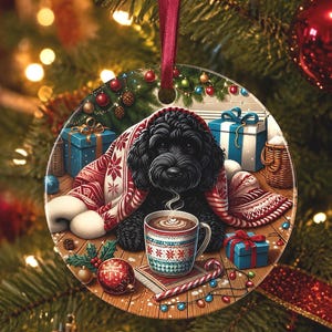May include: A round ceramic Christmas ornament featuring a black curly-haired dog wrapped in a red and white patterned blanket. The dog sits behind a steaming mug of hot cocoa on a wooden surface surrounded by blue gift boxes and a candy cane.