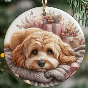 Peut inclure: Ornement de Noël rond en céramique représentant un chien Cavalier King Charles Spaniel. Le chien est allongé sur une couverture en tricot grise. L'arrière-plan comprend un cadeau emballé, des fleurs et une tasse. Une ficelle dorée est attachée.