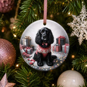 Peut inclure: Une décoration de Noël ronde en céramique représentant un épagneul noir portant un pull à motifs rouges et blancs avec un collier à breloque cœur, entouré de cadeaux emballés et d'une guirlande lumineuse sur un fond gris clair.