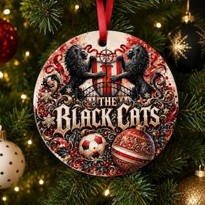 May include: A round ceramic Christmas ornament featuring two black lions flanking a central shield with a sword. Below, the text THE BLACK CATS appears in a bold font, accompanied by a soccer ball and a red patterned bauble with gold accents.